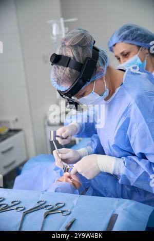 Chirurgien féminin utilise une lampe frontale chirurgicale tout en travaillant, avec un patient sous anesthésie sur la table chirurgicale Banque D'Images