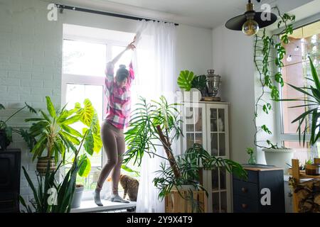 Femme accroche des rideaux de tulle transparents sur de grandes fenêtres dans la maison à l'intérieur. Nettoyage de printemps, rangement Banque D'Images