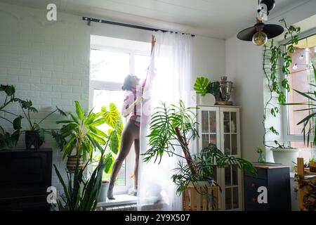 Femme accroche des rideaux de tulle transparents sur de grandes fenêtres dans la maison à l'intérieur. Nettoyage de printemps, rangement Banque D'Images