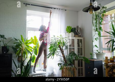 Femme accroche des rideaux de tulle transparents sur de grandes fenêtres dans la maison à l'intérieur. Nettoyage de printemps, rangement Banque D'Images