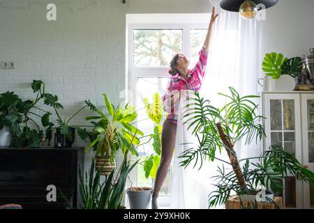 Femme accroche des rideaux de tulle transparents sur de grandes fenêtres dans la maison à l'intérieur. Nettoyage de printemps, rangement Banque D'Images