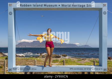 Afrique du Sud, Western Cape, Cape Town, jeune femme faisant une pause dans un cadre photo géant sur Robben Island, site classé au patrimoine mondial de l'UNESCO, vue sur le Cap et le parc national de table Mountain Banque D'Images