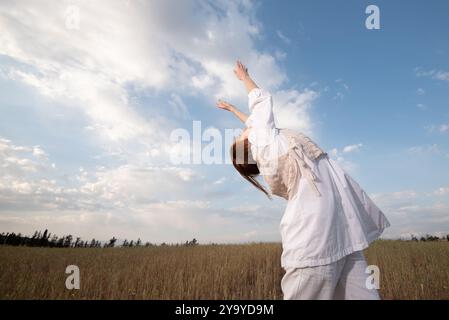 Jeune femme portant une robe blanche dansant à l'extérieur contre un ciel nuageux bleu. Banque D'Images