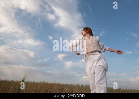 Jeune femme portant une robe blanche dansant à l'extérieur contre un ciel nuageux bleu. Banque D'Images