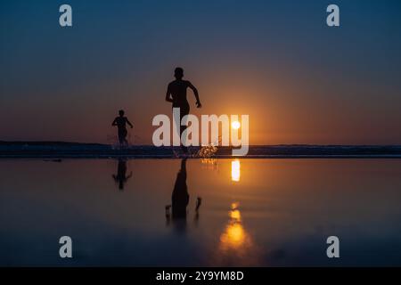 Deux personnages courant le long d'une plage au coucher du soleil, leurs reflets capturés dans l'eau calme alors que le soleil se couche sur l'horizon. Des éclaboussures d'eau se forment en courant Banque D'Images