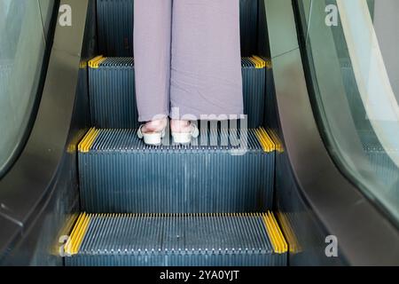 Vue rapprochée des pieds d'une personne sur un escalier roulant. L'individu porte des sandales de couleur claire et un pantalon gris ample. Les escaliers roulants Banque D'Images