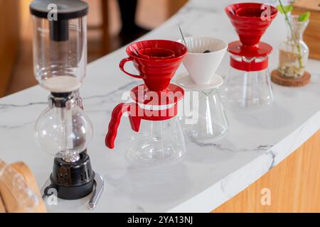 Une installation élégante de préparation du café avec une cafetière rouge à verser, un compte-gouttes en céramique blanche et une cafetière siphon sur un comptoir en marbre. Le Banque D'Images