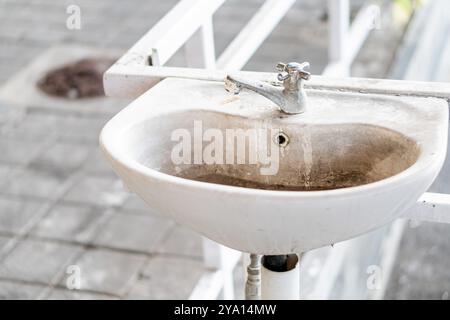 Un vieil évier sale avec de l'eau stagnante, situé dans une zone abandonnée ou négligée. L'évier est blanc avec un robinet rouillé et est partiellement entouré par Banque D'Images