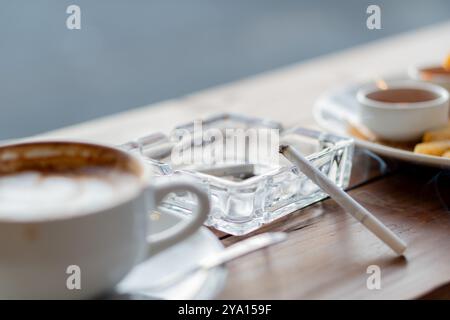 Une scène de café confortable avec une tasse de café, un cendrier avec une cigarette, et une assiette de collations. Le cadre a une atmosphère chaleureuse et accueillante, perfe Banque D'Images