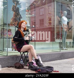 Jeune femme aux cheveux roux assise et jouant à la cornemuse devant une vitrine de magasin affichant des vêtements modernes, Tralee Banque D'Images