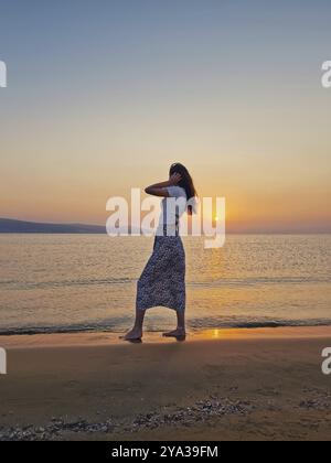 Belle jeune femme marchant pieds nus dans l'eau sur la plage à l'aube, regardant le lever de soleil chaud au-dessus de la mer. Scène matinale sereine bord de mer, summe Banque D'Images