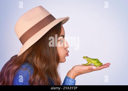 Visage, grenouille et profil de femme s'embrassant en studio sur fond bleu pour fantaisie, espoir ou souhait. Amour, magie ou narration et personne tenant le crapaud dedans Banque D'Images