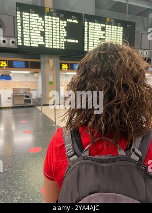 Une jeune femme brune avec les cheveux bouclés est vue de derrière, portant un sac à dos, debout dans un aéroport et regardant le tableau de départ, préparant f Banque D'Images