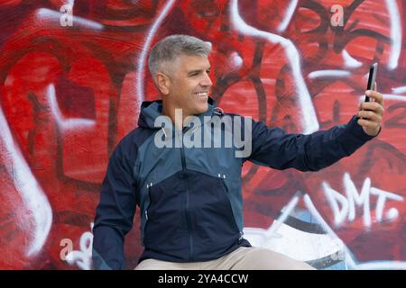 Homme aux cheveux gris dans ses années 40 sourires alors qu'il prend un selfie contre un mur de graffiti vert, portant un imperméable bleu. Atmosphère urbaine et décontractée de style de rue Banque D'Images