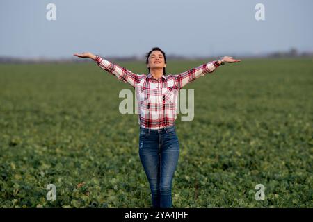 Jolie jeune femme fermière debout avec les mains levées dans un champ vert, se sentant heureuse, excitée et satisfaite Banque D'Images