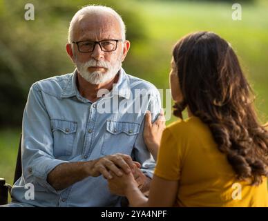 Jeune femme soignante tenant la main d'un homme âgé, communiquant, soutenant et prenant soin des personnes âgées. Assis à l'extérieur dans le parc avec verdure Banque D'Images