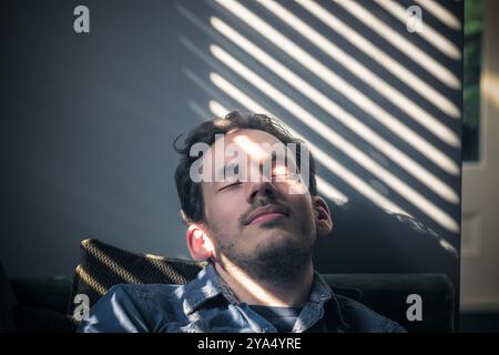 Portrait d'un homme caucasien somnolant sur le canapé, sur le visage tombe l'ombre des stores Banque D'Images