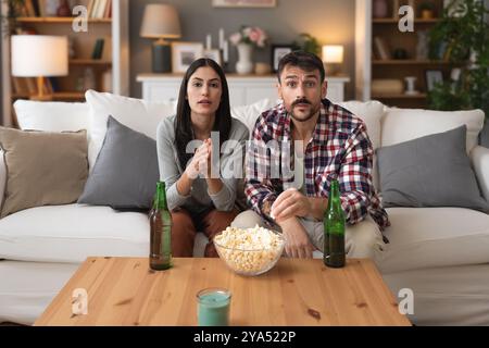 Jeune homme de couple et femme assis à la maison sur le canapé prenant une pause après une journée de travail stressante regarder un jeu de sport ou un film à la télévision, manger du pop-corn et du drin Banque D'Images