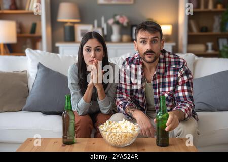 Jeune homme de couple et femme assis à la maison sur le canapé prenant une pause après une journée de travail stressante regarder un jeu de sport ou un film à la télévision, manger du pop-corn et du drin Banque D'Images