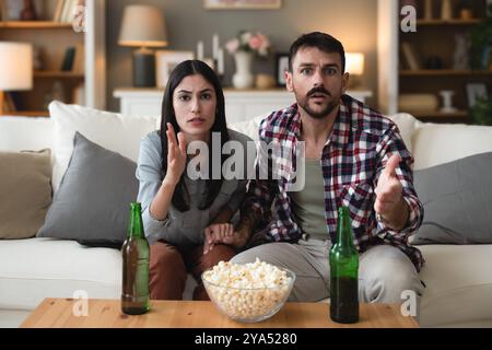 Jeune homme de couple et femme assis à la maison sur le canapé prenant une pause après une journée de travail stressante regarder un jeu de sport ou un film à la télévision, manger du pop-corn et du drin Banque D'Images