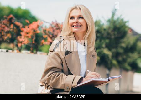 Photo de belle femme rêveuse habillée dans des vêtements élégants profitant de la journée ensoleillée d'automne écrivant des notes dans le journal Banque D'Images