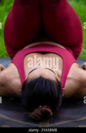 Tête de jeune femme au premier plan soulevant les jambes pour effectuer une pose de yoga habillée de leggings rouges confortables et top avec la nature verte en arrière-plan Banque D'Images