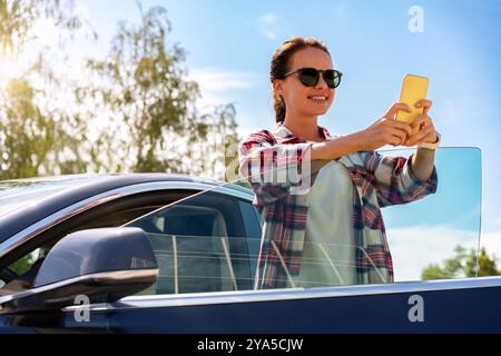 Femme debout à côté de sa voiture à l'extérieur, textant sur son téléphone portable. Banque D'Images