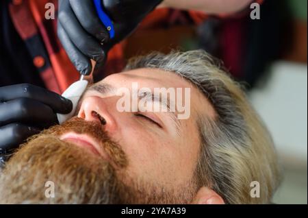 Soin professionnel de la barbe. Homme barbu dans un salon de coiffure. Coiffeur servant le client au salon de coiffure. Barbe homme. Rasoir droit, salon de coiffure. Outils de coiffure. V Banque D'Images