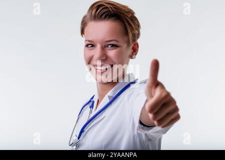 Médecin féminin confiant sourit et donne un pouce vers le haut, symbolisant l'approbation dans les soins de santé. Elle porte un manteau blanc avec un stéthoscope Banque D'Images
