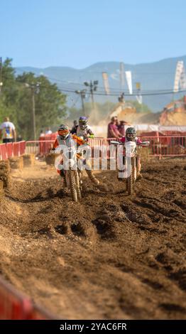 Les coureurs de motocross courent sur une piste boueuse lors d'une compétition de circuit de montagne en plein air avec des gens regardant depuis les clôtures. Banque D'Images