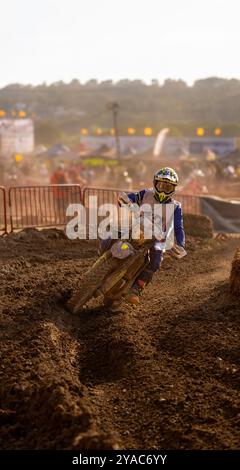 Un coureur de motocross se pliant dans un virage serré sur une piste de terre humide éclairée par la lumière du soleil du soir sur un circuit extérieur de motocross et d'enduro avec PA Banque D'Images