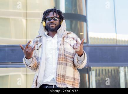 Drôle d'homme africain avec des lunettes de soleil, des dreadlocks et des écouteurs dansant dans une ville. émotions positives Banque D'Images