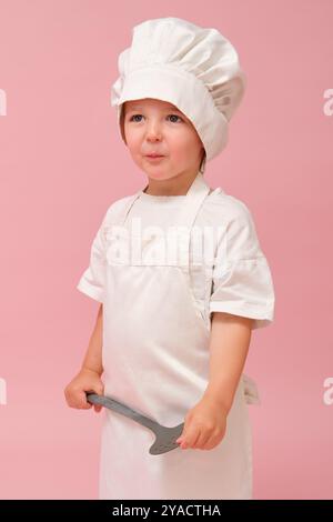 Un jeune garçon dans un chapeau de chef et un tablier tient des ustensiles de cuisine et fait un visage drôle sur un fond rose dans le studio. Enfant âgé de 3 ans (t Banque D'Images