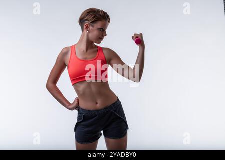 Femme confiante fléchissant bicep avec haltère rose dans le studio de gymnastique, mettant en valeur la détermination et la force. Athlète en forme respire la confiance dans la vie active Banque D'Images