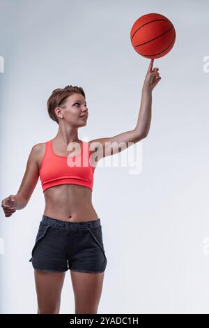 Fit jeune femme habilement tourne un ballon de basket sur le bout de son doigt, exsudant confiance et détermination dans les vêtements de sport sur un fond blanc propre Banque D'Images