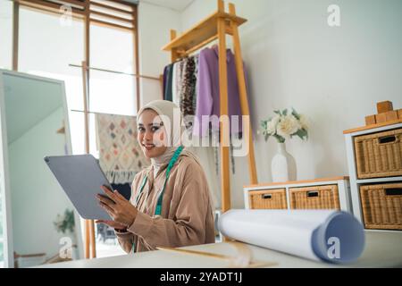 Belle créatrice de mode asiatique voilée utilise une tablette dans l'atelier Banque D'Images