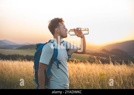 Randonneur avec un sac à dos bleu boit dans une bouteille d'eau tout en se tenant debout dans un champ ensoleillé au coucher du soleil. La lumière dorée, les collines lointaines, et surrou paisible Banque D'Images