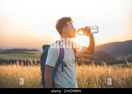 Randonneur avec un sac à dos bleu boit dans une bouteille d'eau tout en se tenant debout dans un champ ensoleillé au coucher du soleil. La lumière dorée, les collines lointaines, et surrou paisible Banque D'Images