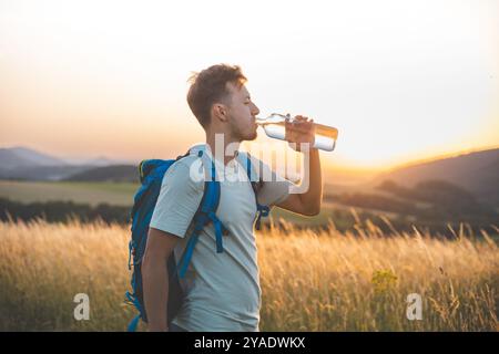 Randonneur avec un sac à dos bleu boit dans une bouteille d'eau tout en se tenant debout dans un champ ensoleillé au coucher du soleil. La lumière dorée, les collines lointaines, et surrou paisible Banque D'Images