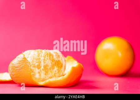 Deux oranges sur un fond rose vif, une orange est partiellement pelée, mettant en valeur son intérieur juteux. Les couleurs vives créent un contraste saisissant, Banque D'Images