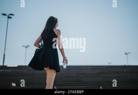 Vue arrière d'une femme dans une robe noire tenant une bouteille d'eau marchant dans les escaliers à l'extérieur Banque D'Images