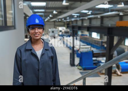 Jeune ingénieur confiant, une femme indienne, dans un casque bleu et un manteau de travail gris dans une usine, portrait photographié. Banque D'Images