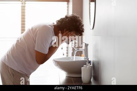 Un jeune indien se lavant le visage avec de l'eau claire du robinet se pliant au-dessus de l'évier, faisant la routine de soin du visage dans la salle de bains Banque D'Images