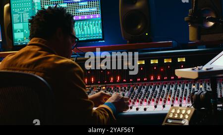 Ingénieur audio afro-américain utilisant la console de mixage sonore pour la gestion des pistes, la post-production musicale. Producteur appuyant sur les boutons de commande motorisés, regarde le logiciel d'édition. Caméra A. Banque D'Images