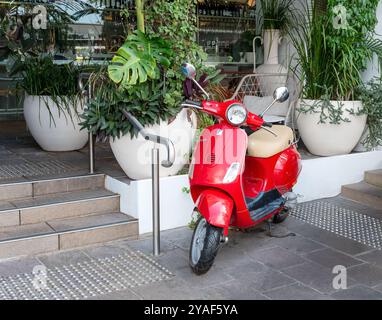 BRISBANE, QUEENSLAND, AUSTRALIE. 11 septembre 2024 ; scooter Vespa rouge garé devant un bar décoré de feuillage vert. Banque D'Images