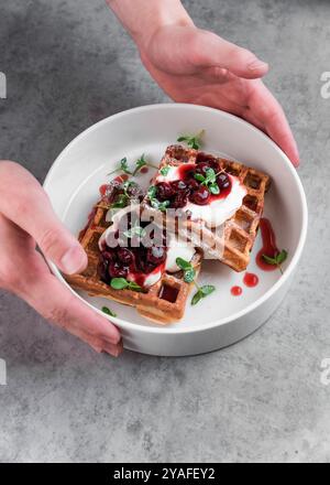 Serveur servant une assiette blanche avec des gaufres belges sucrées avec de la crème et de la confiture de cerises sur une table grise dans un café Banque D'Images