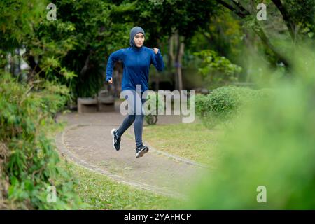 Femme musulmane mignonne asiatique portant des vêtements de sport et un hijab bleu s'exerçant et courant à l'extérieur au parc le matin. avec une sensation de fraîcheur. Banque D'Images