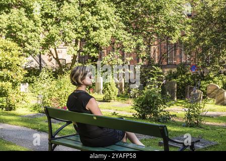 Femme de trente ans dans une robe noire assise sur un banc dans un cimetière en été, Suède, Europe Banque D'Images