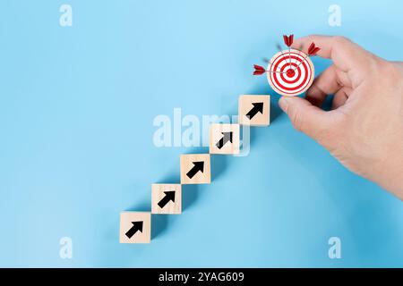 Main d'homme tenant le bloc en bois avec le tableau de fléchettes de visée de cible et flèche vers le haut sur fond bleu. Réalisation de l'entreprise, objectif objectif et avancement du projet Banque D'Images