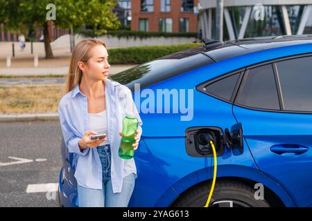 Femme se tient à côté d'une voiture électrique bleue en charge, tenant un verre et vérifiant son téléphone Banque D'Images